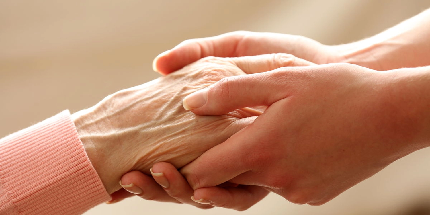 Eine Krankenpflegerin hält die alten Hände einer pflegebedürftigen Person um sie zu trösten.