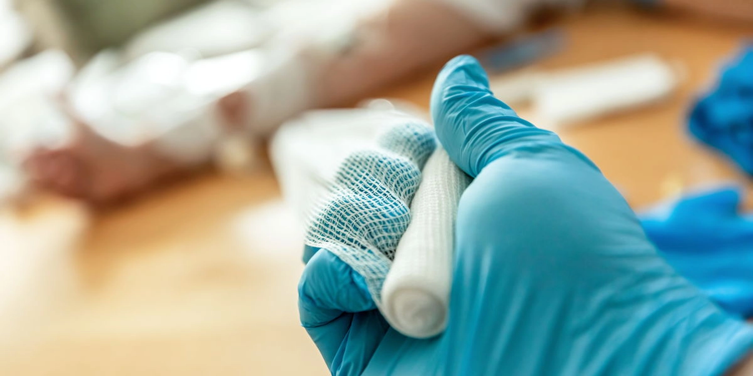 Eine Pflegerin mit blauen Latexhandschuhen hält eine Mullbinde in der Hand, um eine hygienische Wundbehandlung sicher zu stellen.